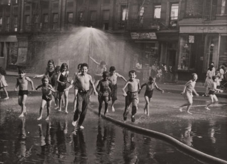 black and white photograph of children on city street playing under a fire house spraying water in the air