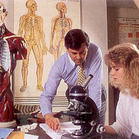 woman seated at microscope with professor leaning over, anatomical drawings in background