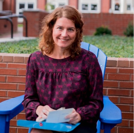 head shot of Laurie Mathes smiling and seated on an Adirondack chair in front of the Abplanalp Library.